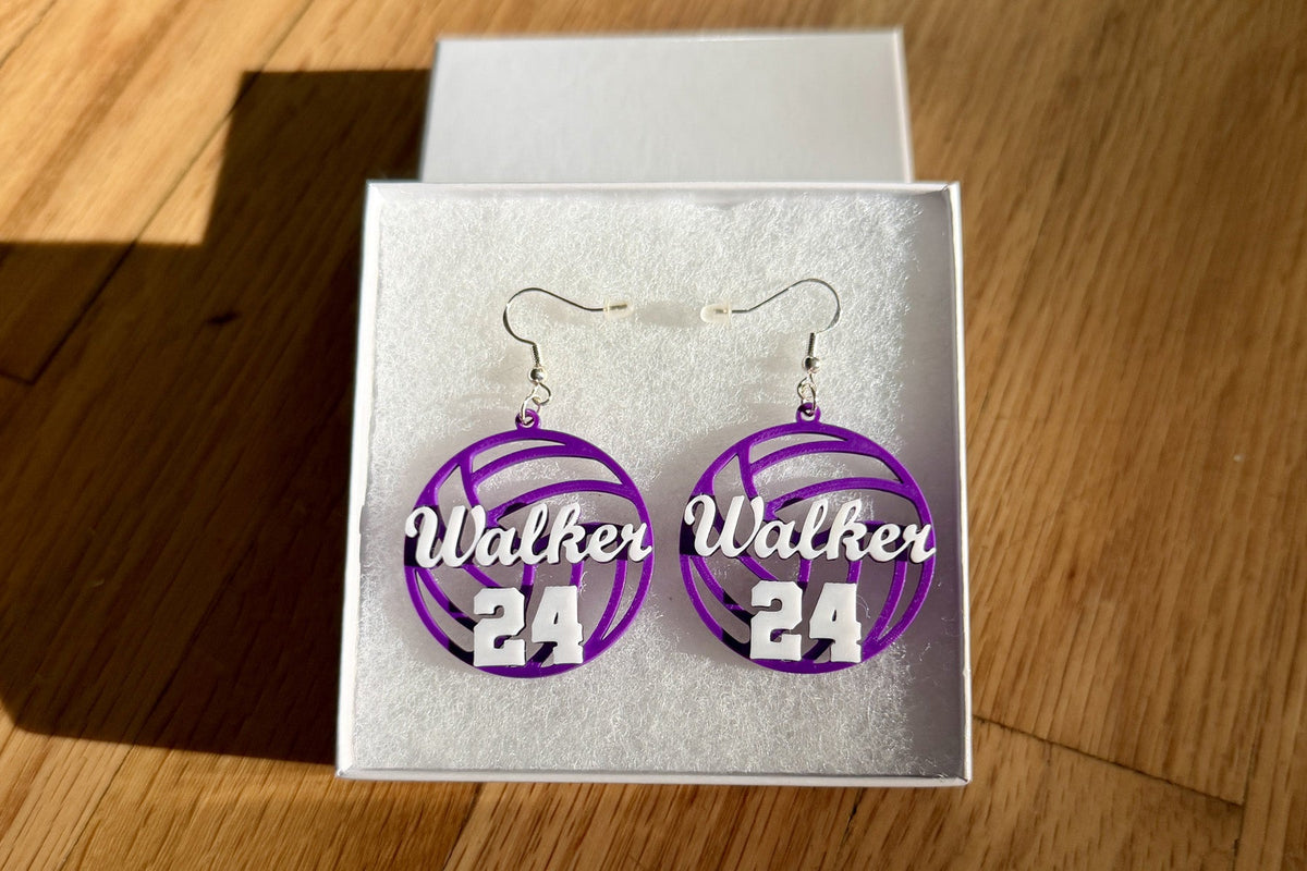 Purple and white personalized volleyball earrings with Walker and number 24 displayed in a white jewelry box on a wooden surface.