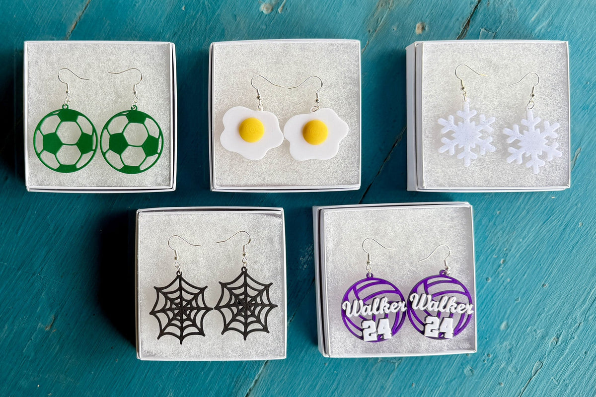 Collection of Hook & Charm earrings including green soccer balls, fried eggs, white snowflakes, black spider webs, and personalized purple volleyball earrings displayed in jewelry boxes.