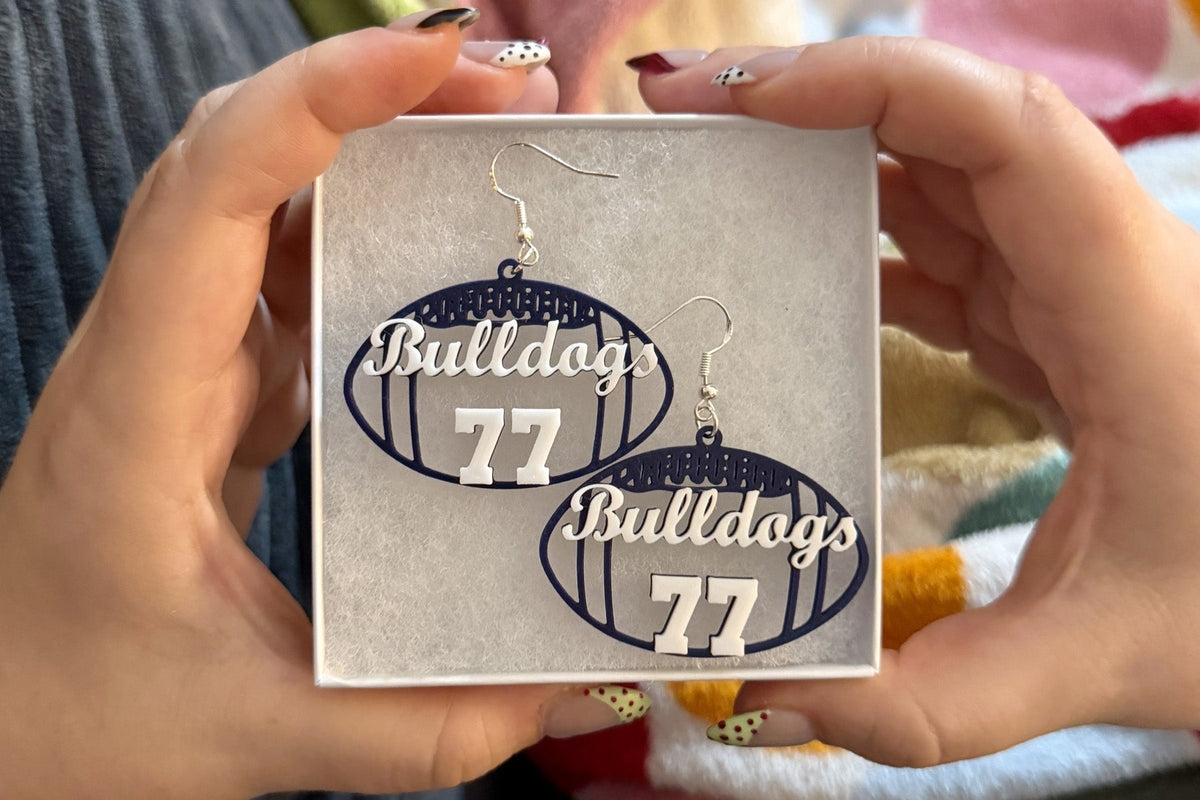 Navy and white personalized football earrings with Bulldogs and number 77 displayed in a jewelry box held in both hands.
