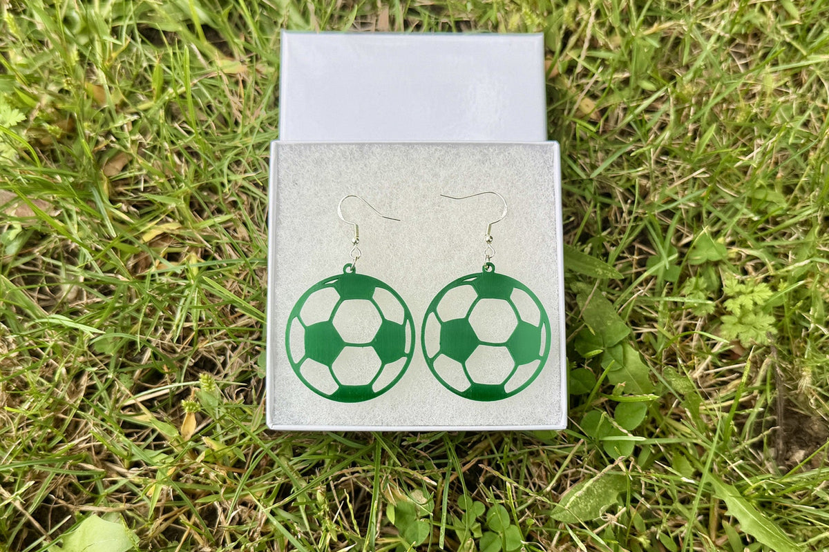 Collection of sports earrings featuring softball, soccer, volleyball, basketball, football helmet, football and baseball designs displayed on grass.