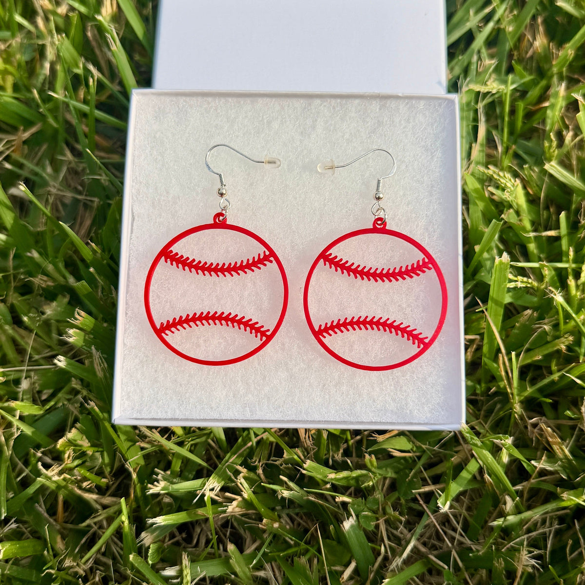 Pair of red baseball earrings with open stitching design displayed in a jewelry box on grass.