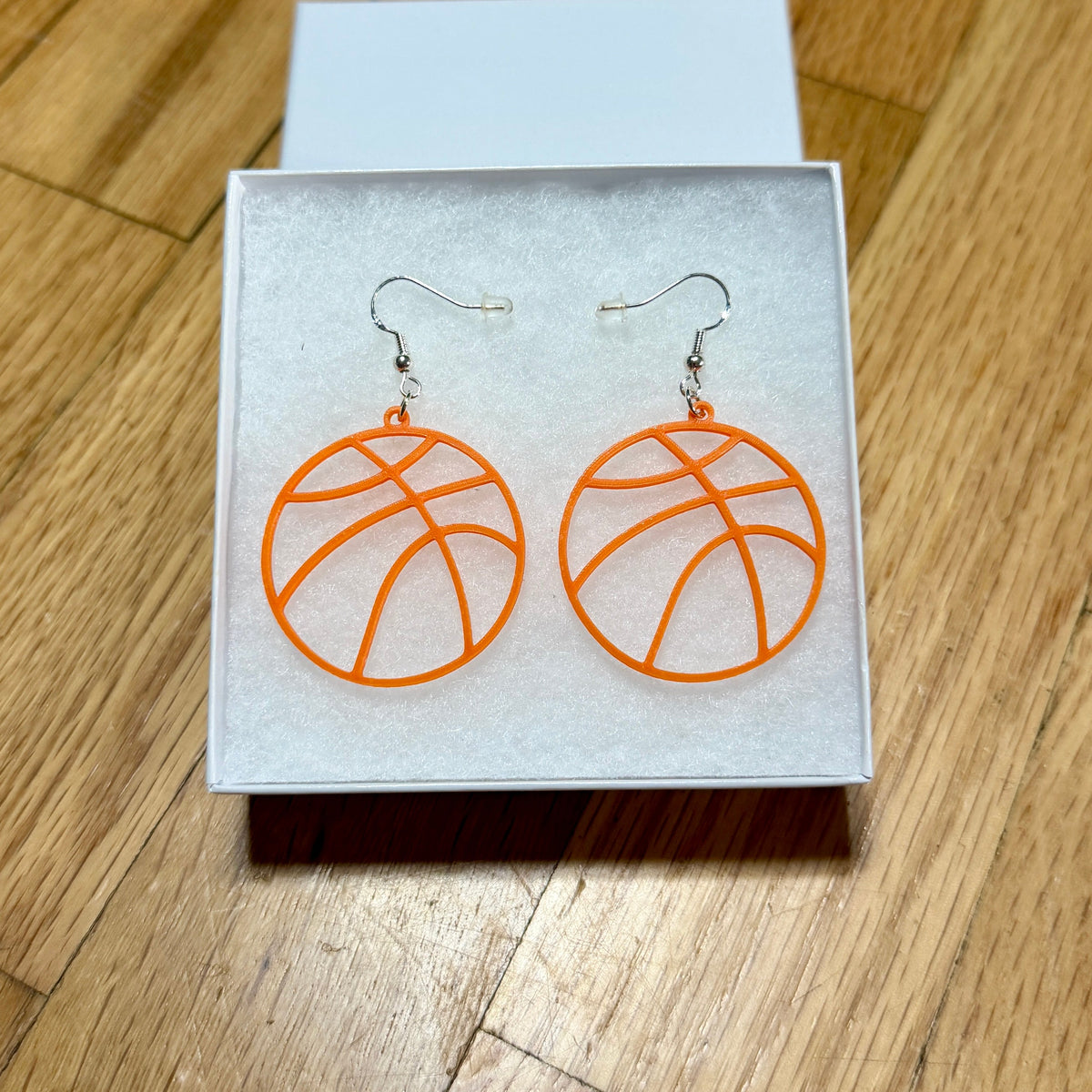 Pair of orange basketball earrings with open line design displayed in a jewelry box.