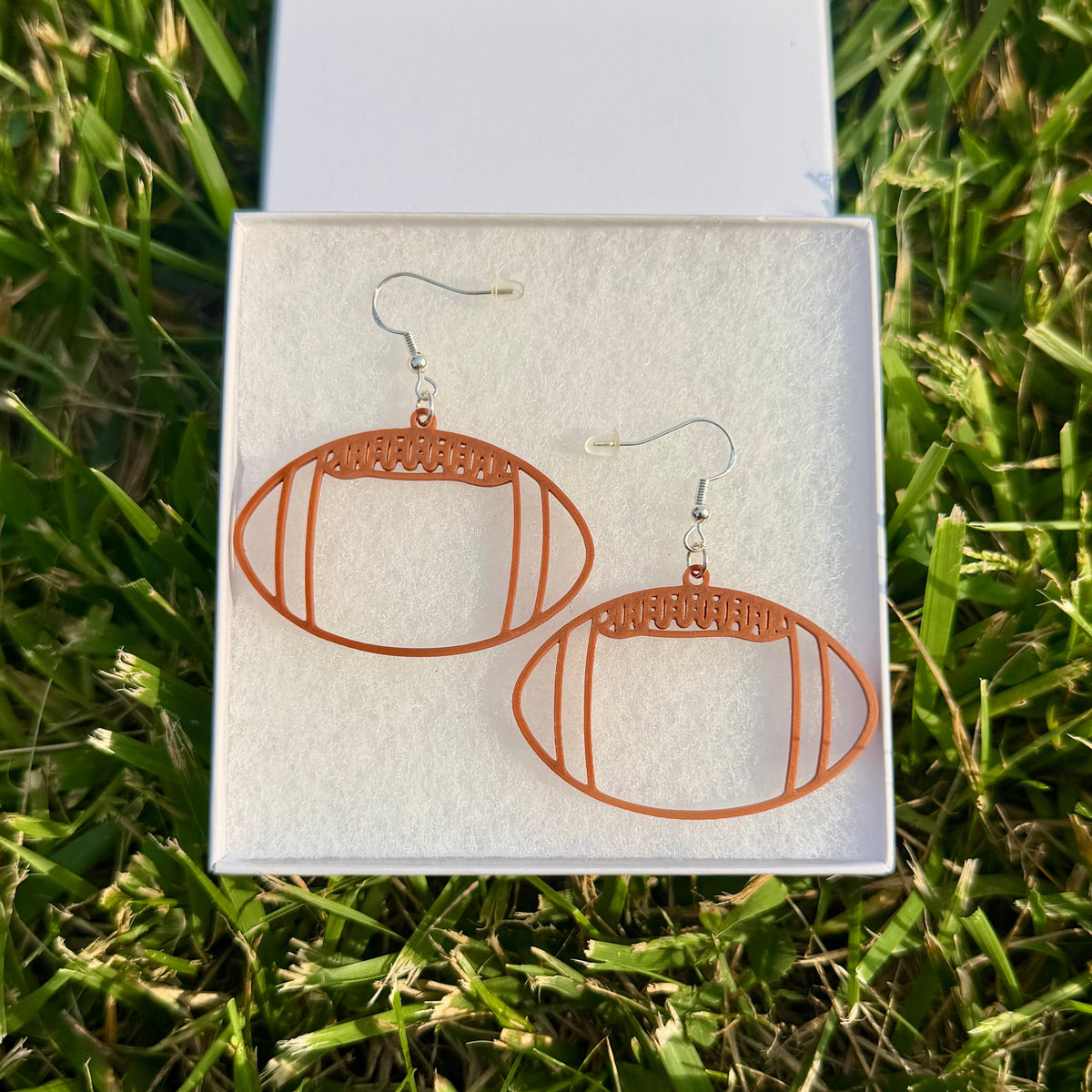 Pair of brown football earrings with open line design displayed in a jewelry box on grass.
