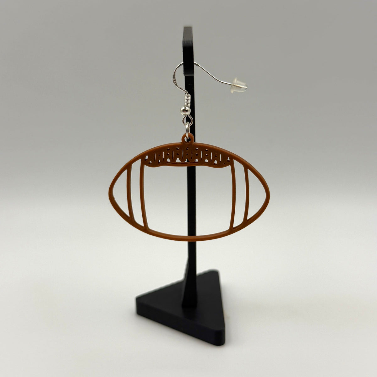 Brown football earring with open line design displayed on a black stand.