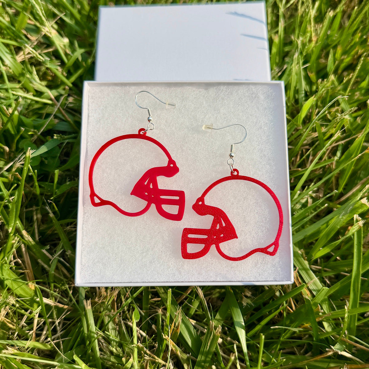 Pair of red football helmet earrings with open design displayed in a jewelry box on grass.