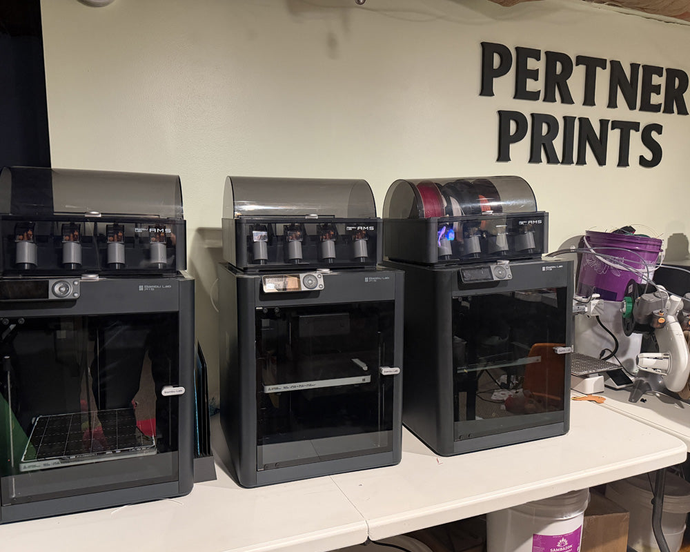 Row of Bambu Lab 3D printers and filament systems in the Hook & Charm production workspace.