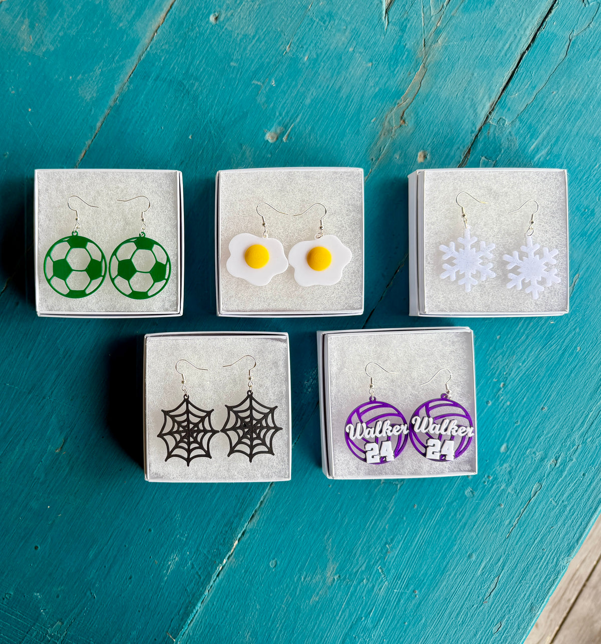 Collection of Hook & Charm earrings including green soccer balls, fried eggs, white snowflakes, black spider webs, and personalized purple volleyball earrings displayed in jewelry boxes.