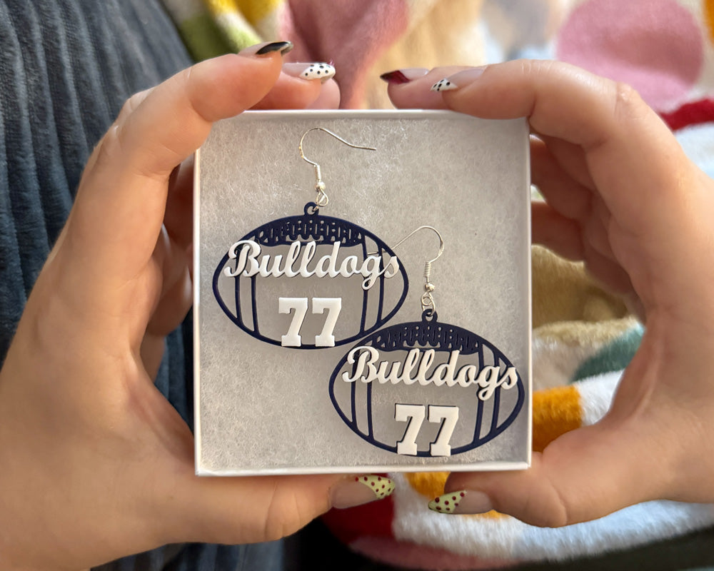 Personalized navy and white football earrings with “Bulldogs” and number 77 displayed in a jewelry box held in both hands.