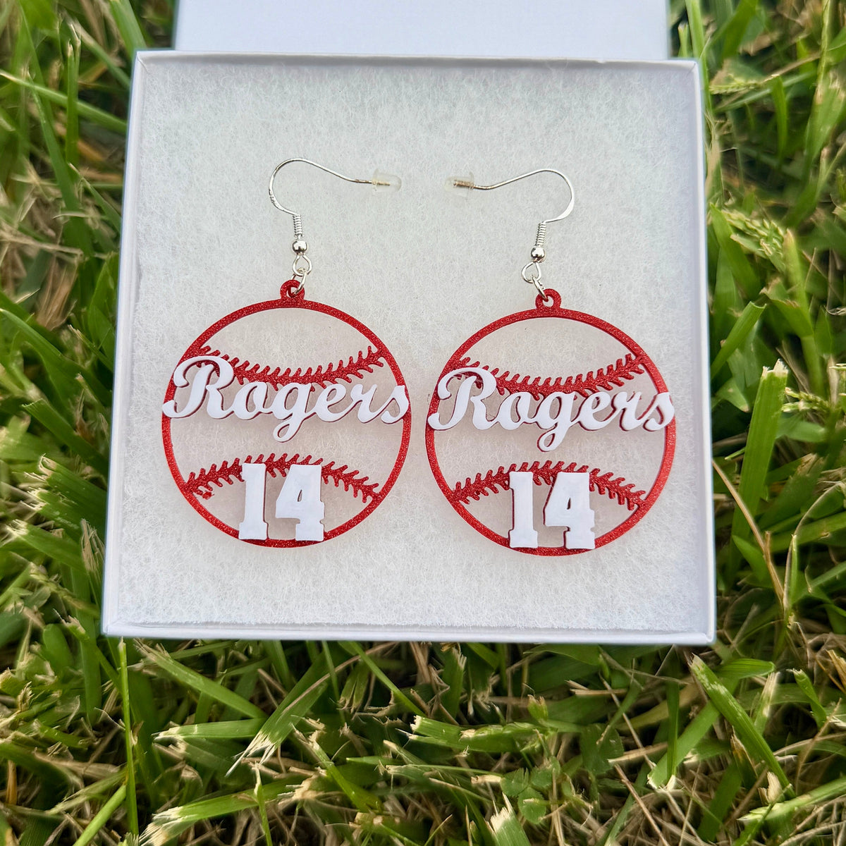 Pair of red and white personalized baseball earrings with Rogers and number 14 displayed in a jewelry box on grass.