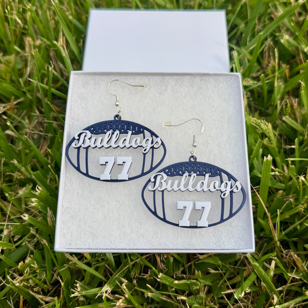 Pair of blue and white personalized football earrings with Bulldogs and number 77 displayed in a jewelry box on grass.