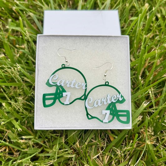 Pair of green and white personalized football helmet earrings with Carter and number 7 displayed in a jewelry box on grass.