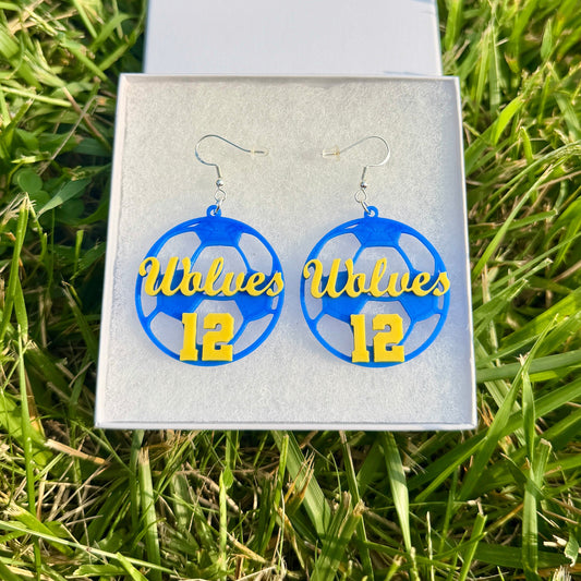 Pair of blue and yellow personalized soccer earrings with Wolves and number 12 displayed in a jewelry box on grass.