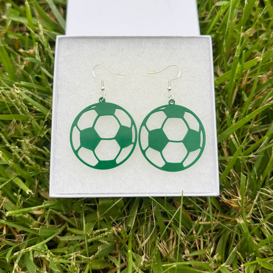 Pair of green soccer ball earrings with open panel design displayed in a jewelry box on grass.
