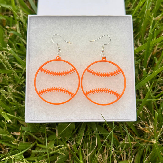 Pair of orange softball earrings with open stitching design displayed in a jewelry box on grass.