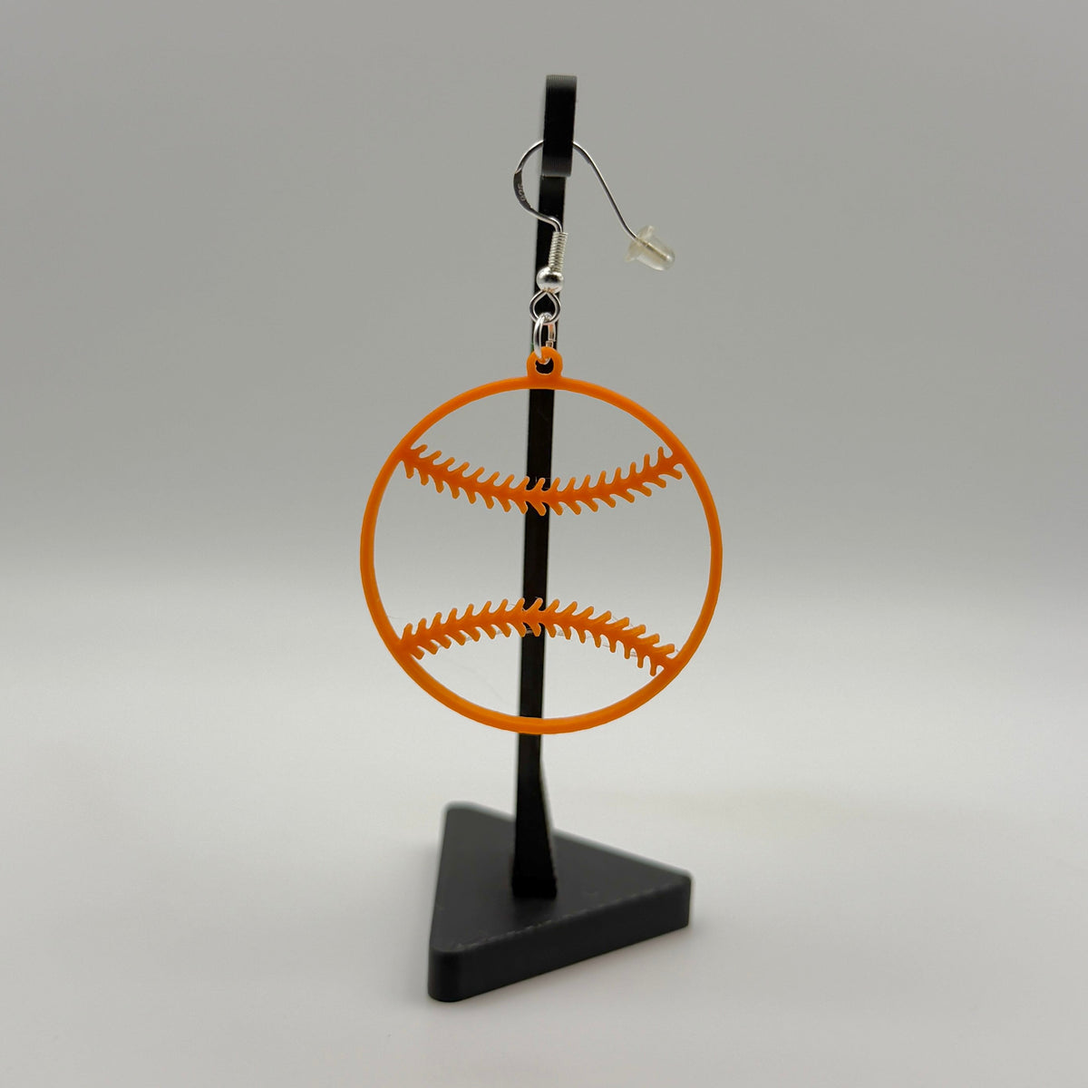 Orange softball earring with open stitching design displayed on a black stand.
