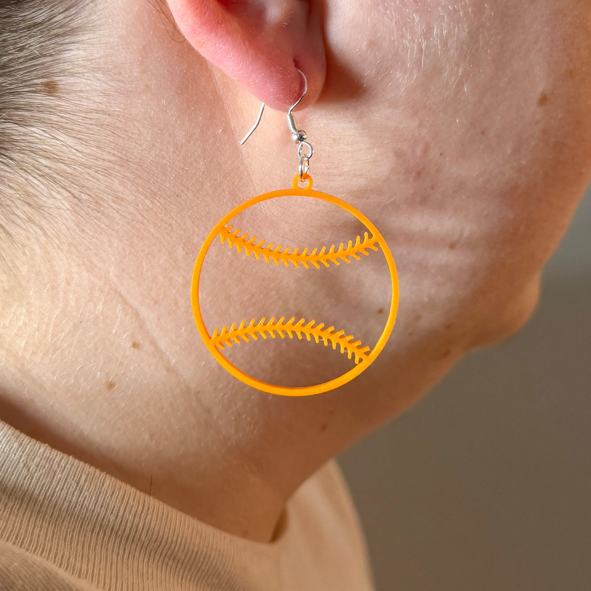Orange softball earring with open stitching design being worn.